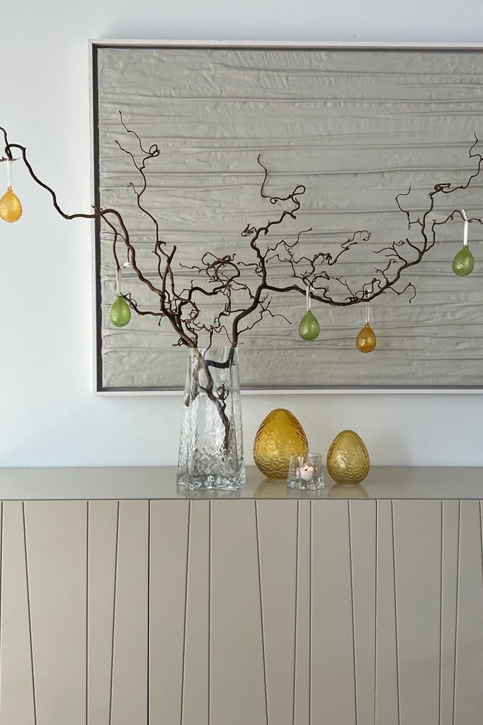 En påskris står på en byrå i vasen Gry, dekorerad med Gry påskhängen, bredvid Gry glasägg, allt från Cooee Design.