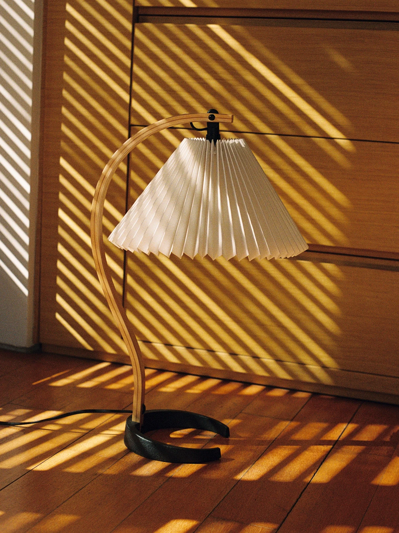 Skrivbordslampa med böjd träfot och veckad skärm, belyst av diagonala solstrålar på ett trägolv.