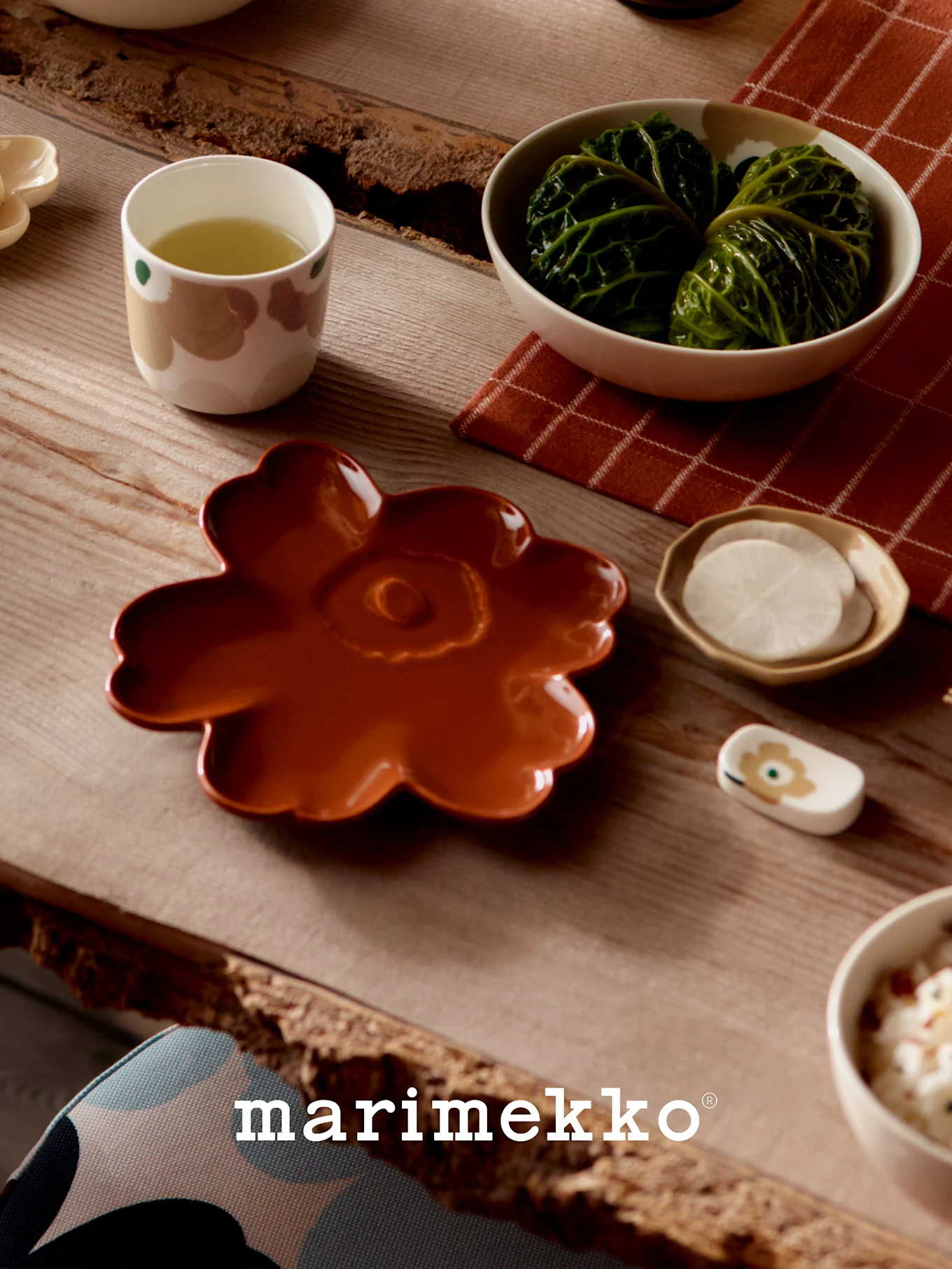 Bord dukat med Marimekko-servis: en rostfärgad blomtallrik, en kopp grönt te, kålrullar och skivade rädisor.
