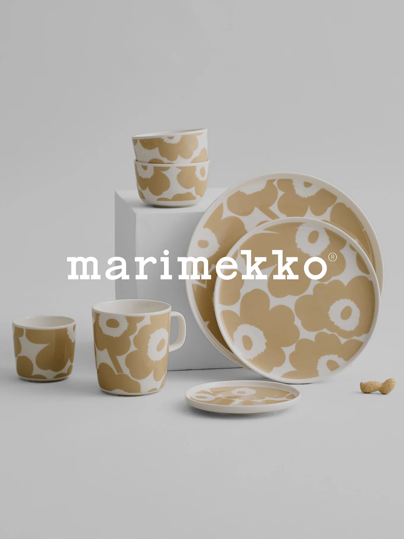 Marimekko middagsservis med beige och vitt blommönster, med skålar, mugg, koppar, tallrikar och en jordnöt på en grå yta.