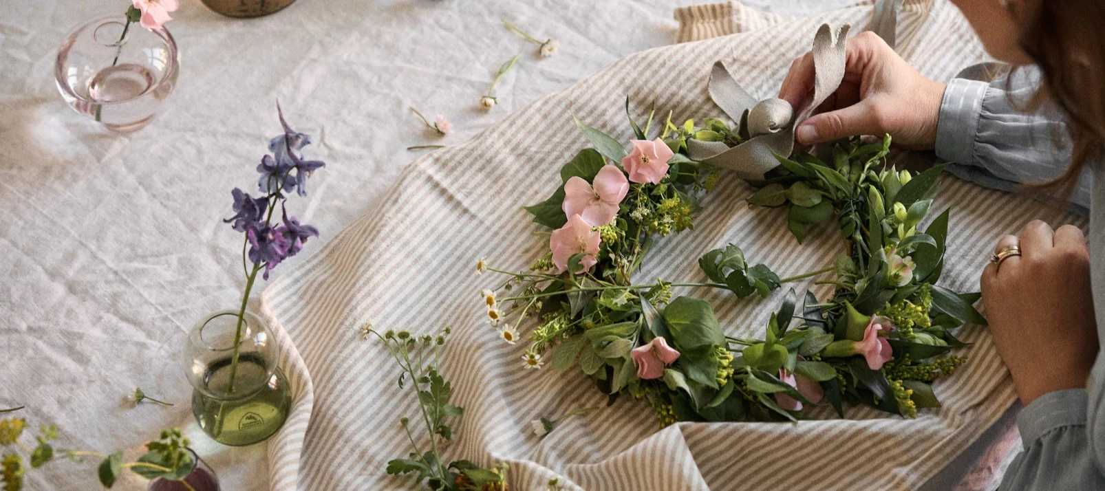 En persons händer fäster ett band vid en blomsterkrans gjord av gröna blad och rosa och vita blommor, placerad på randigt tyg.