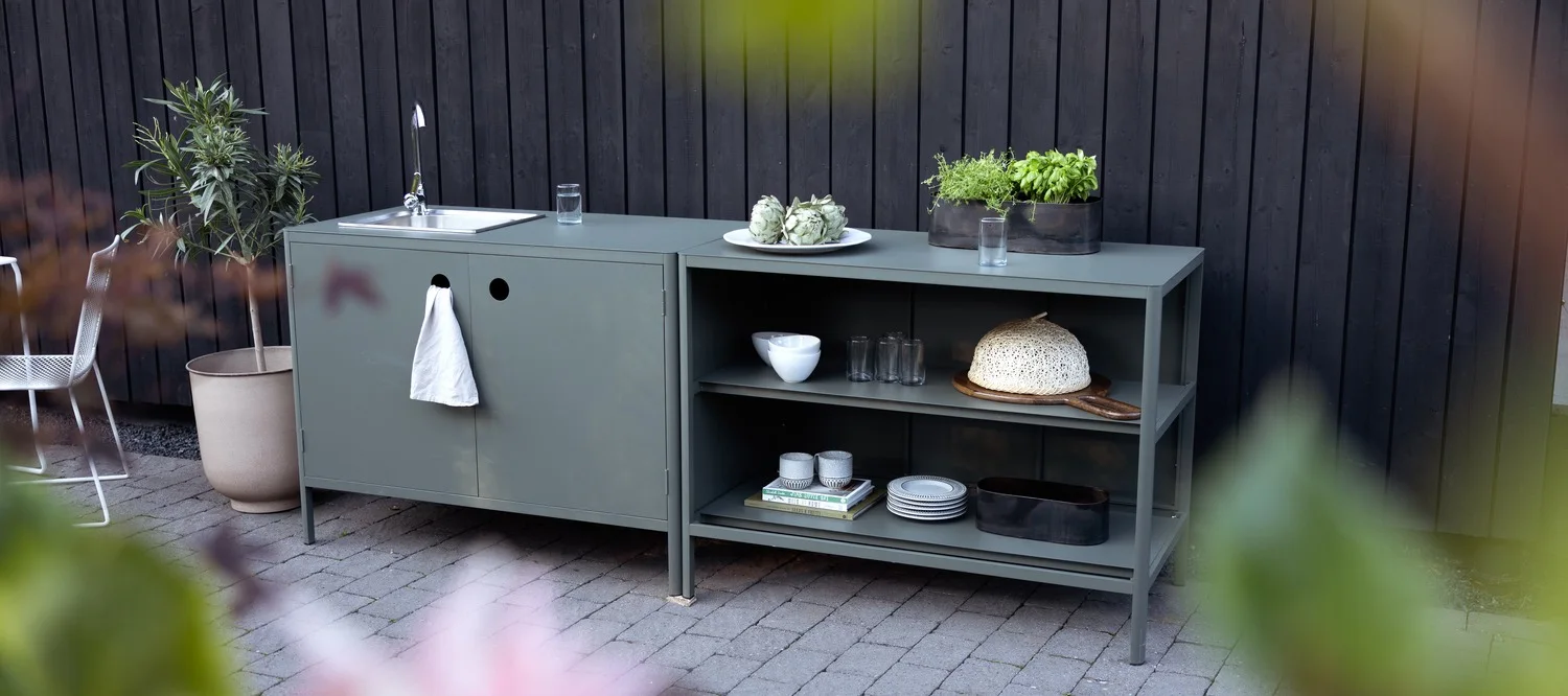 En grågrön utomhusköksmodul, med diskho och handduk till vänster, samt hyllor som visar köksredskap och krukor med örter till höger.
