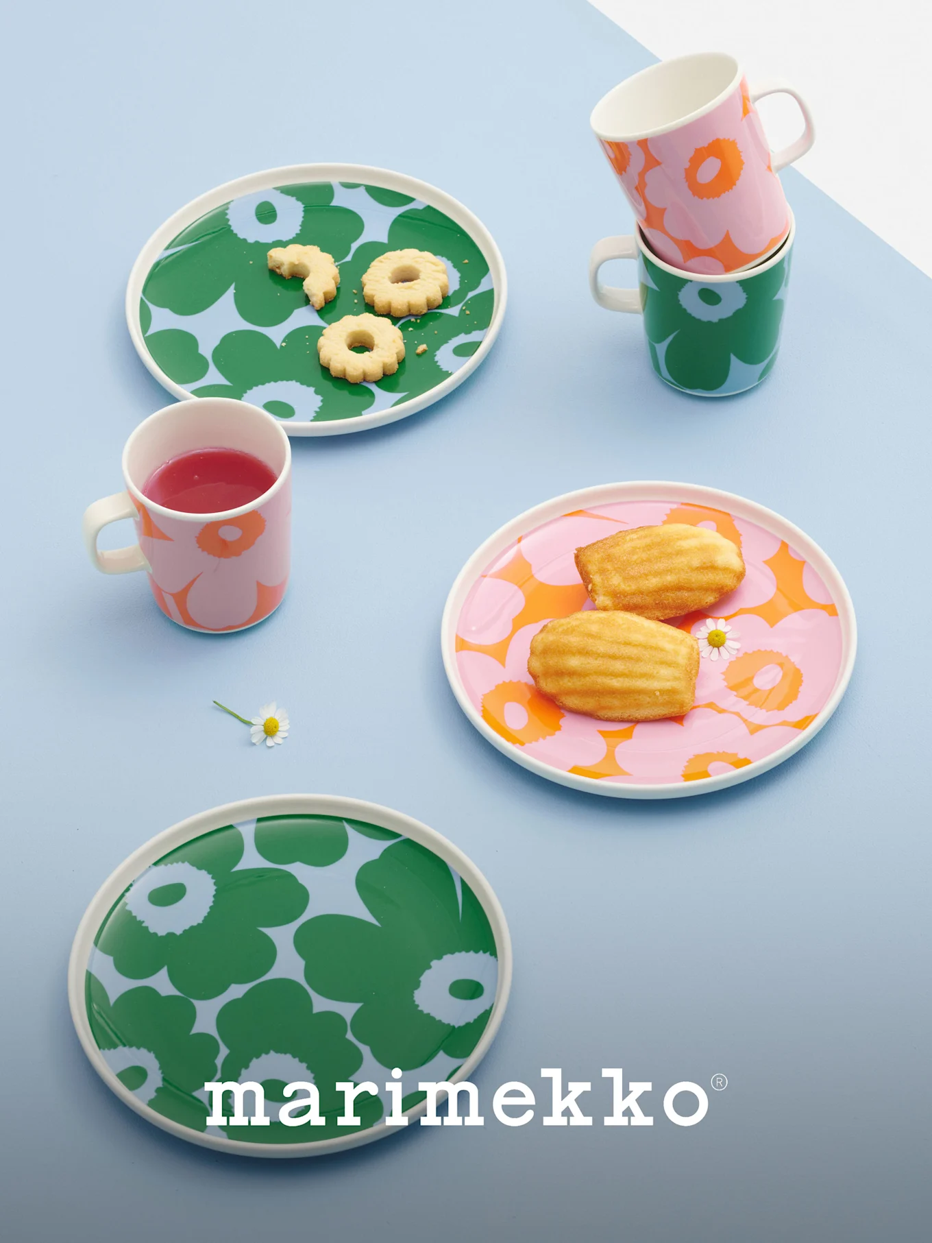 Marimekko tallrikar och muggar med djärva blommönster i grönt, rosa och orange, med kakor, madeleines och en dryck.