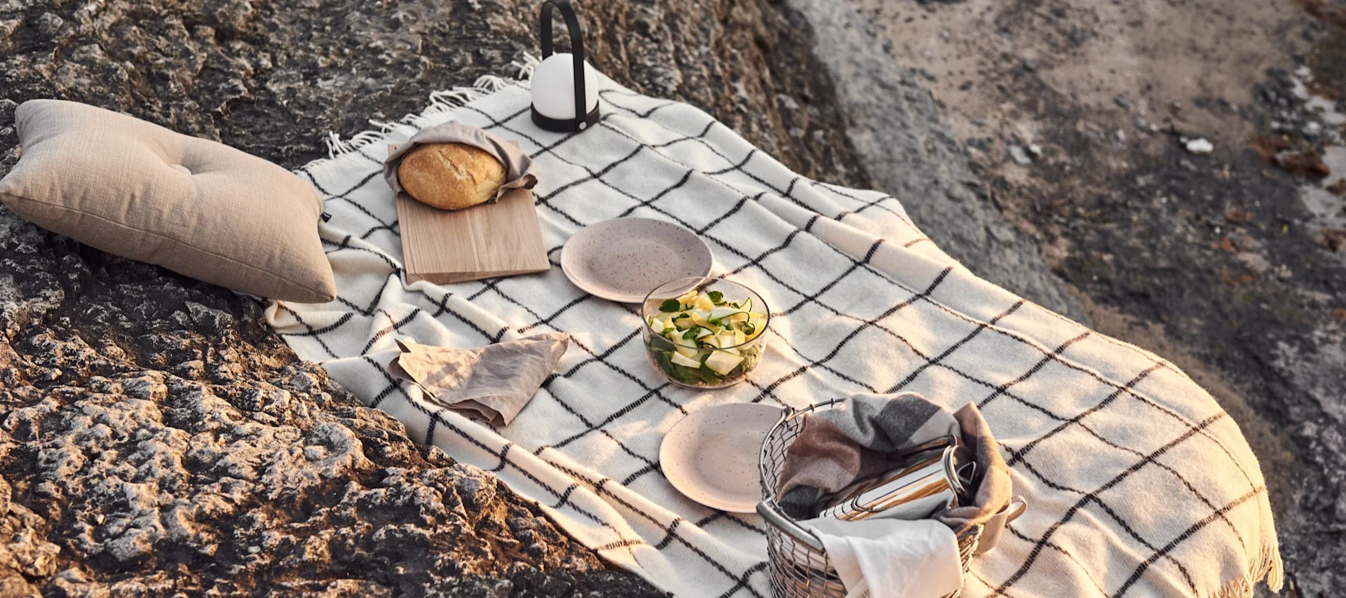 Ett picknickset på karga klippor, med en vitrutig filt, en kudde, bröd, en salladsskål, tallrikar, en lykta och en korg med en filt.