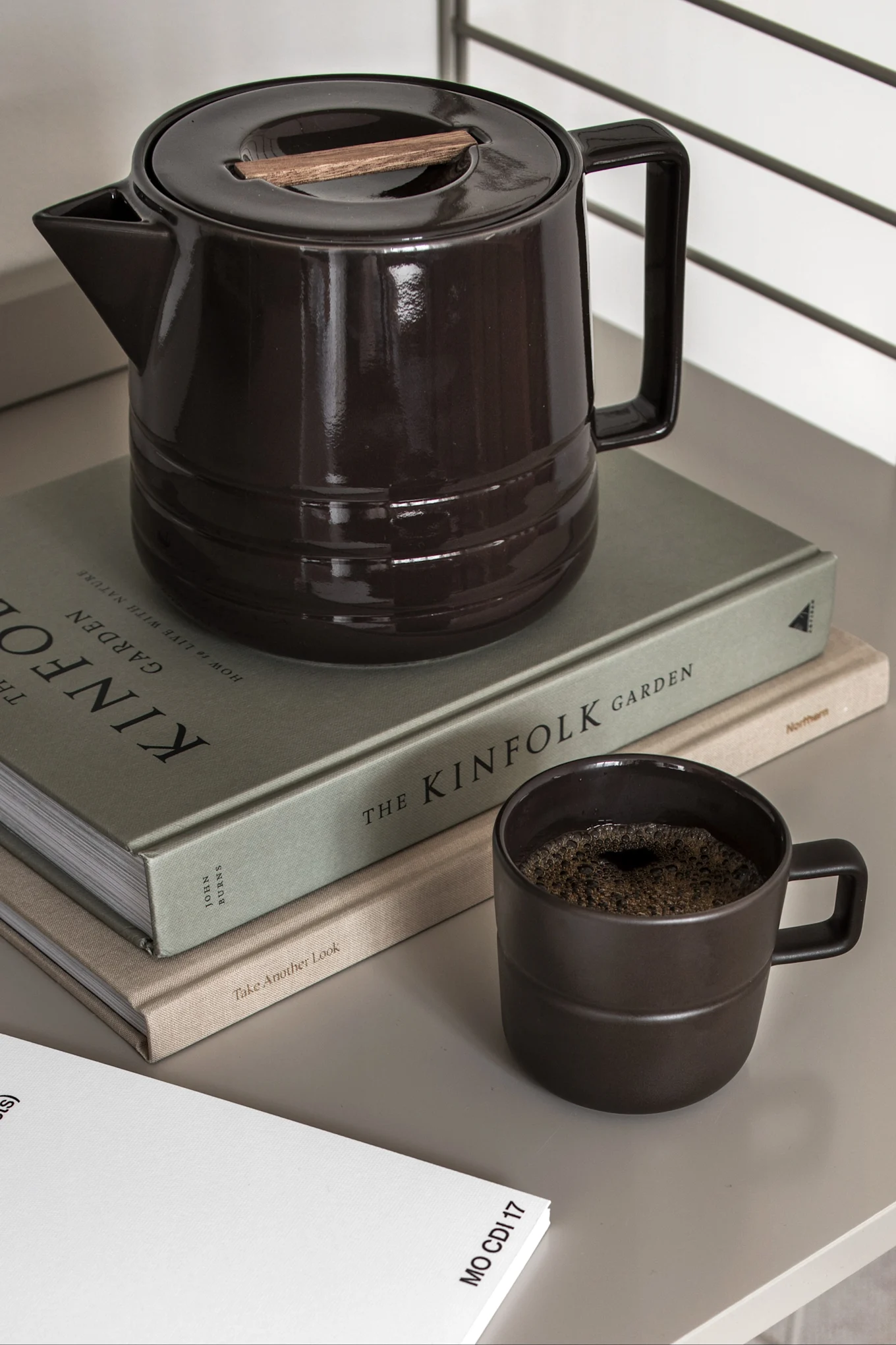 Mörkbrun keramisk kaffekanna med trädetalj på locket, matchande mugg med kaffe och två böcker.