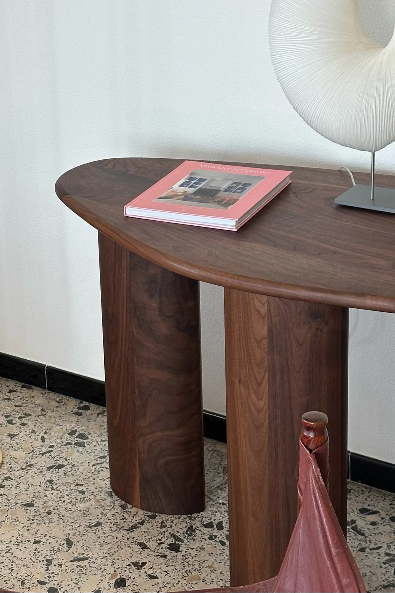 Modernt konsolbord i mörkt trä med en rosa bok och en veckad vit lampa på ett terrazzogolv.