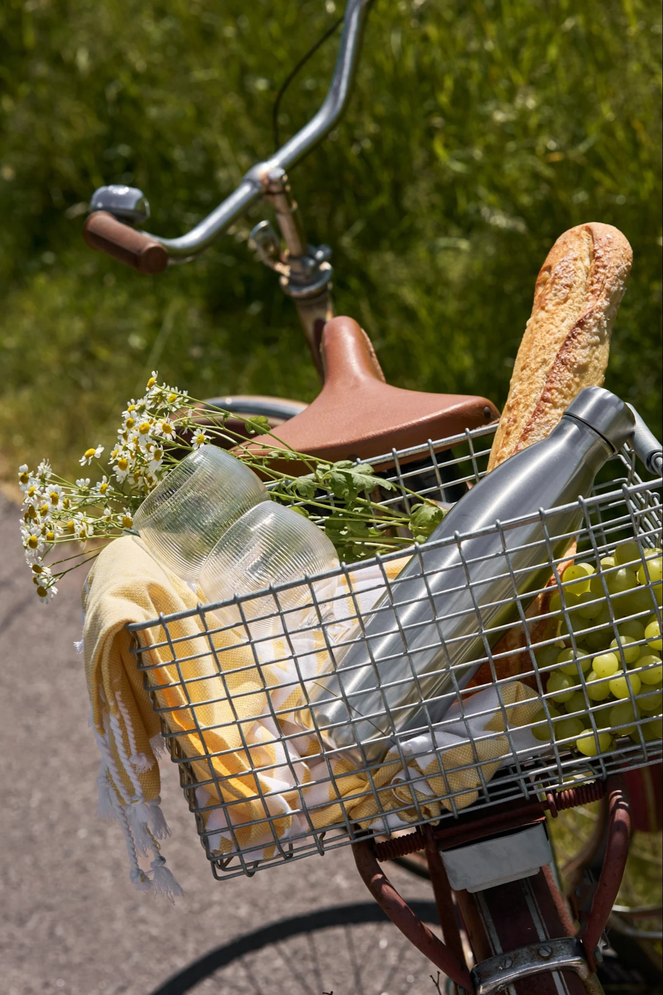 En cykelkorg fylld med picknickartiklar: baguette, vattenflaska, vindruvor, glas, gul handduk och vita prästkragar.