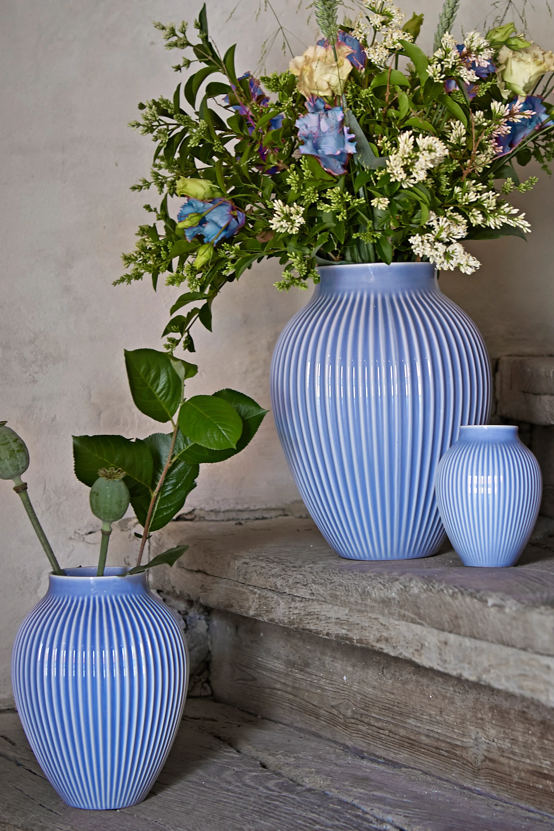 Knabstrup vas i räfflat mönster från Knabstrup keramik står i tre olika storlekar på en stentrappa, alla i samma lavendelblåa färg. Två av vaserna har blomster i sig.