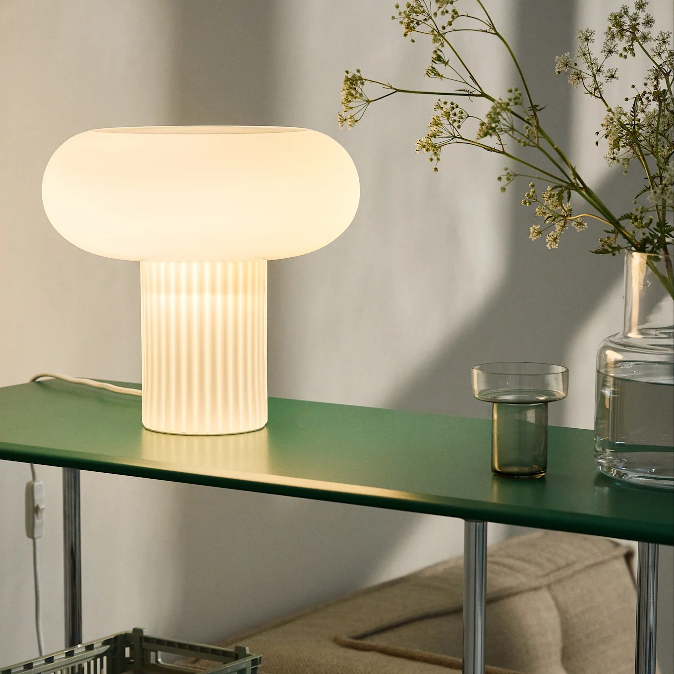 Glödande svampformad bordslampa med räfflad fot på en mörkgrön hylla, tillsammans med blommor i en vas.