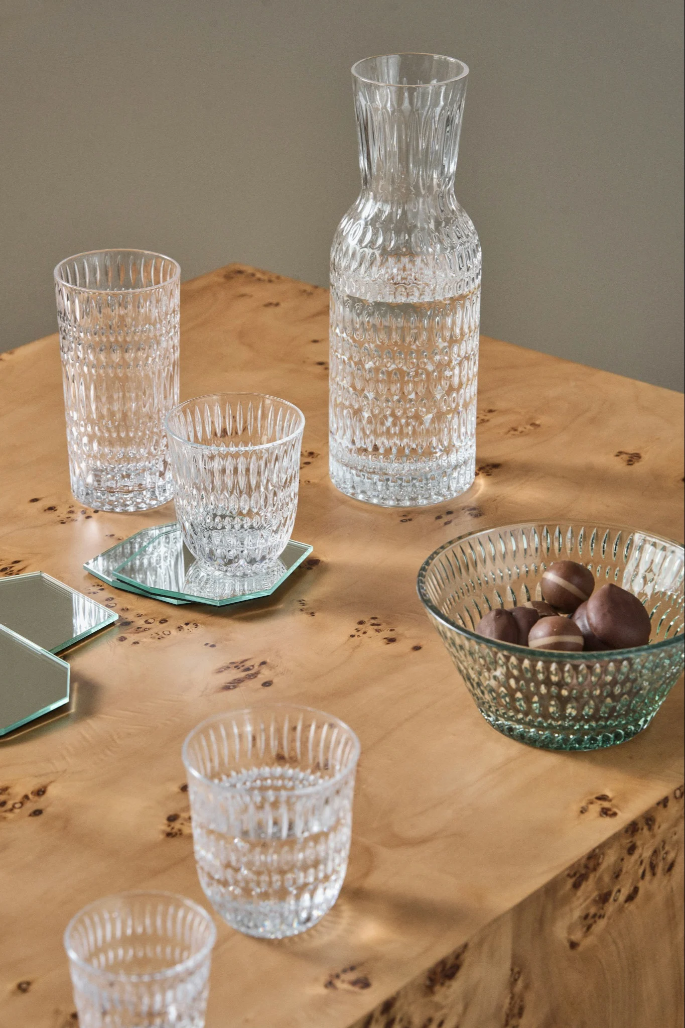 En klar mönstrad glaskaraff, flera glas och en skål choklad på ett ådrat träbord med spegelunderlägg.