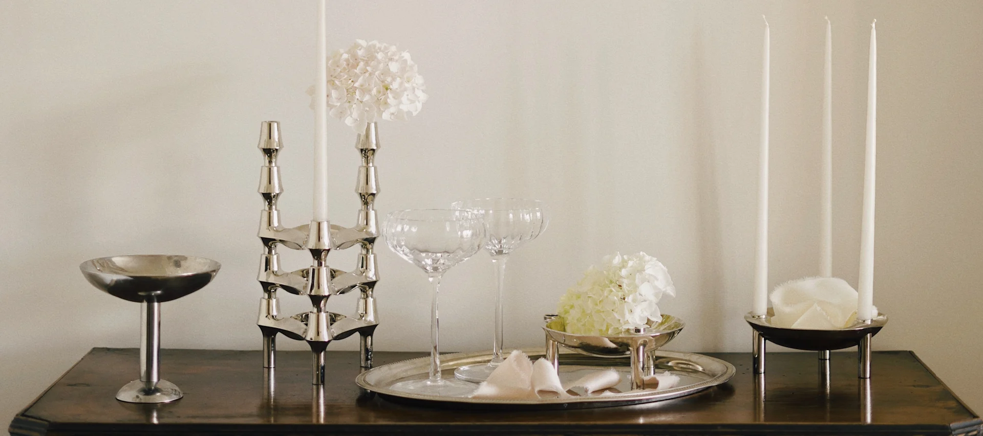 Elegant bordsdekoration med silverljusstakar, vita ljus, glaskupor och vita hortensior på träyta.