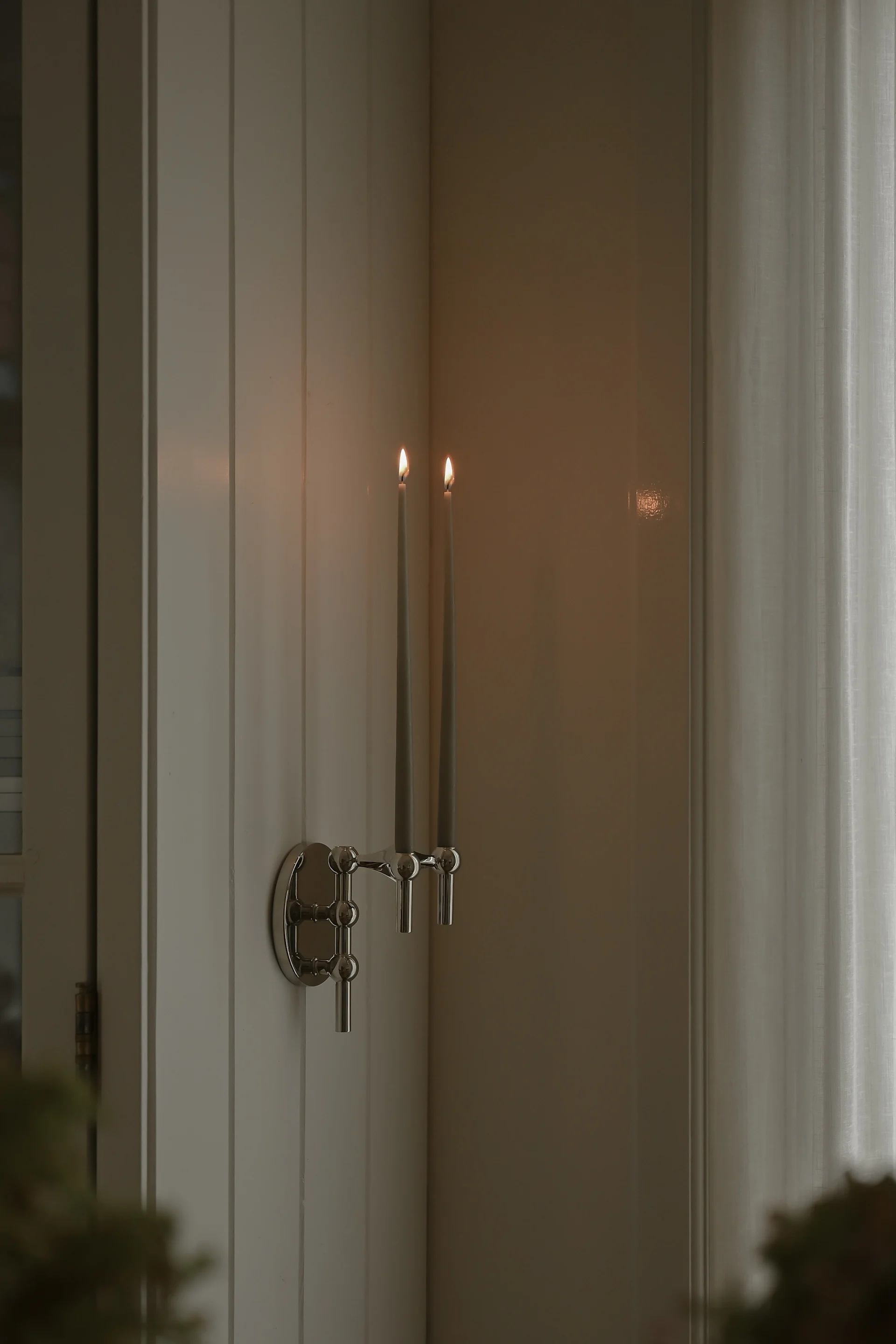 Ett silverfärgat väggmonterat kandelaber håller två tända grå smala ljus på en panelvägg bredvid en skir gardin.