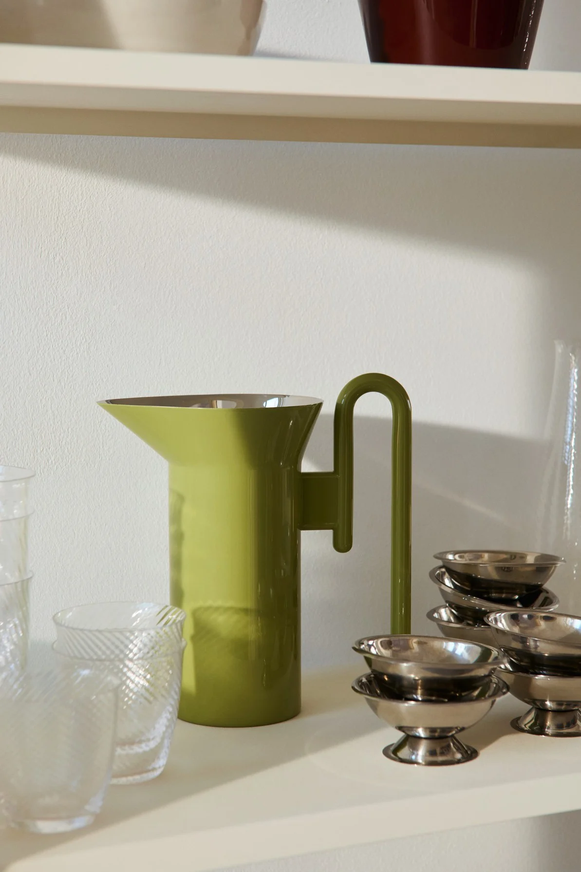 Olivgrön kanna med U-format handtag, flankerad av klara mönstrade glas och staplar med små silverdessertskålar på en hylla.