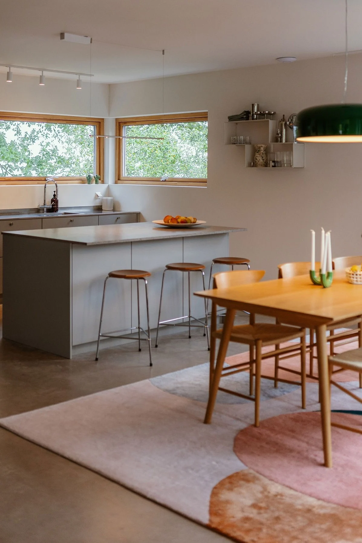 Öppen kök- och matplats med grå ö, tre pallar, ett matbord i trä och en stor grön taklampa.