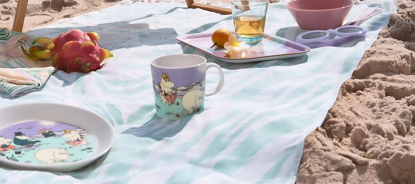 Somrig picknick på stranden med färgglada muminmuggar från Moomin Arabia, brickor och tallrikar med motiv från Beach Day 2025. Muminfamiljen njuter av sol, frukt och havsbris i ett lekfullt dukat strandlandskap.
