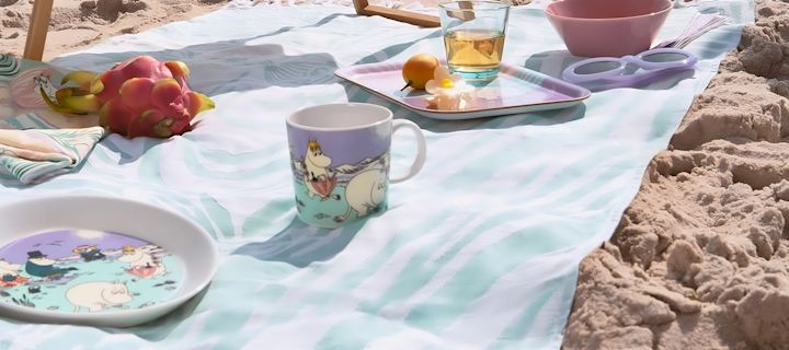 Somrig picknick på stranden med färgglada muminmuggar från Moomin Arabia, brickor och tallrikar med motiv från Beach Day 2025. Muminfamiljen njuter av sol, frukt och havsbris i ett lekfullt dukat strandlandskap.