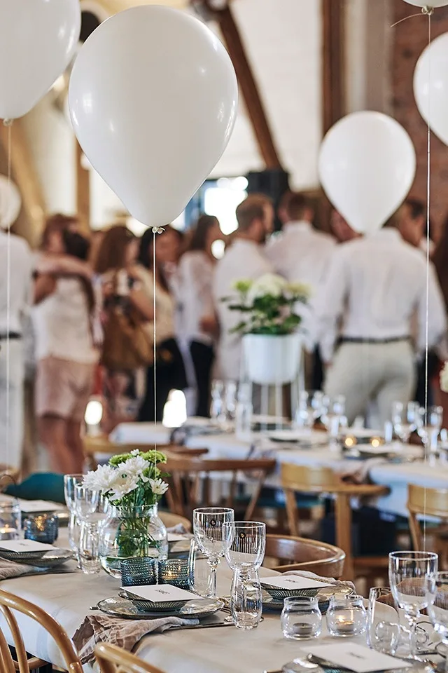 Långbord står uppdukade för sommarfest i vitt och det är dukat för många medan ballonger svävar ovanför borden.