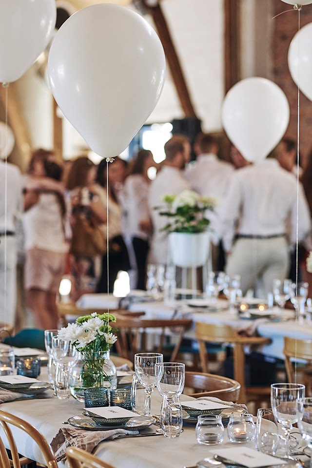 Långbord står uppdukade för sommarfest i vitt och det är dukat för många medan ballonger svävar ovanför borden. 