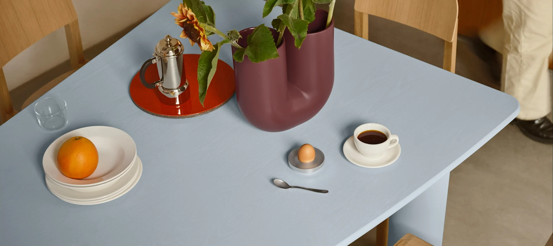 Ljusblått bord dukat med frukost: apelsin på tallrikar, kaffe, ägg i äggkopp och en vas med solrosor.