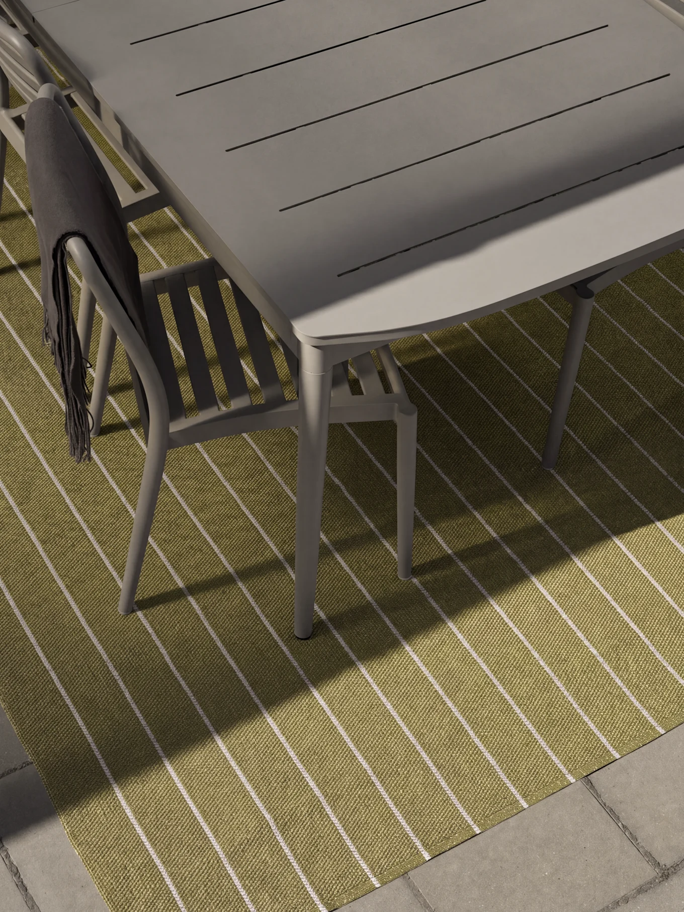 Grått utomhusmatbord och stolar på en grön- och vitrandig matta. En mörkgrå filt ligger draperad över en stol.