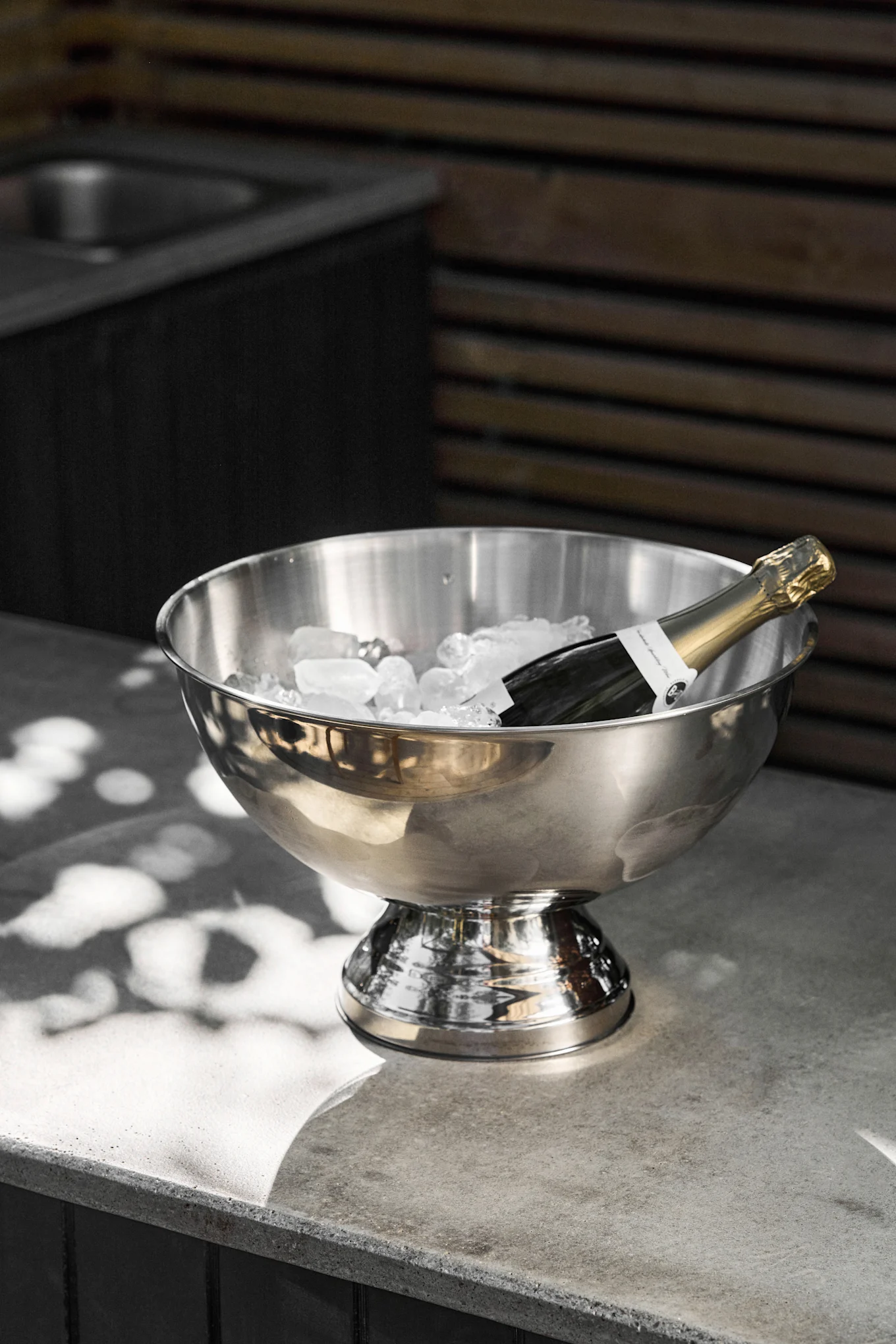 En champagneflaska ligger i en ishink i rostfritt stål fylld med isbitar på en grå betongbänk, med fläckigt solljus.
