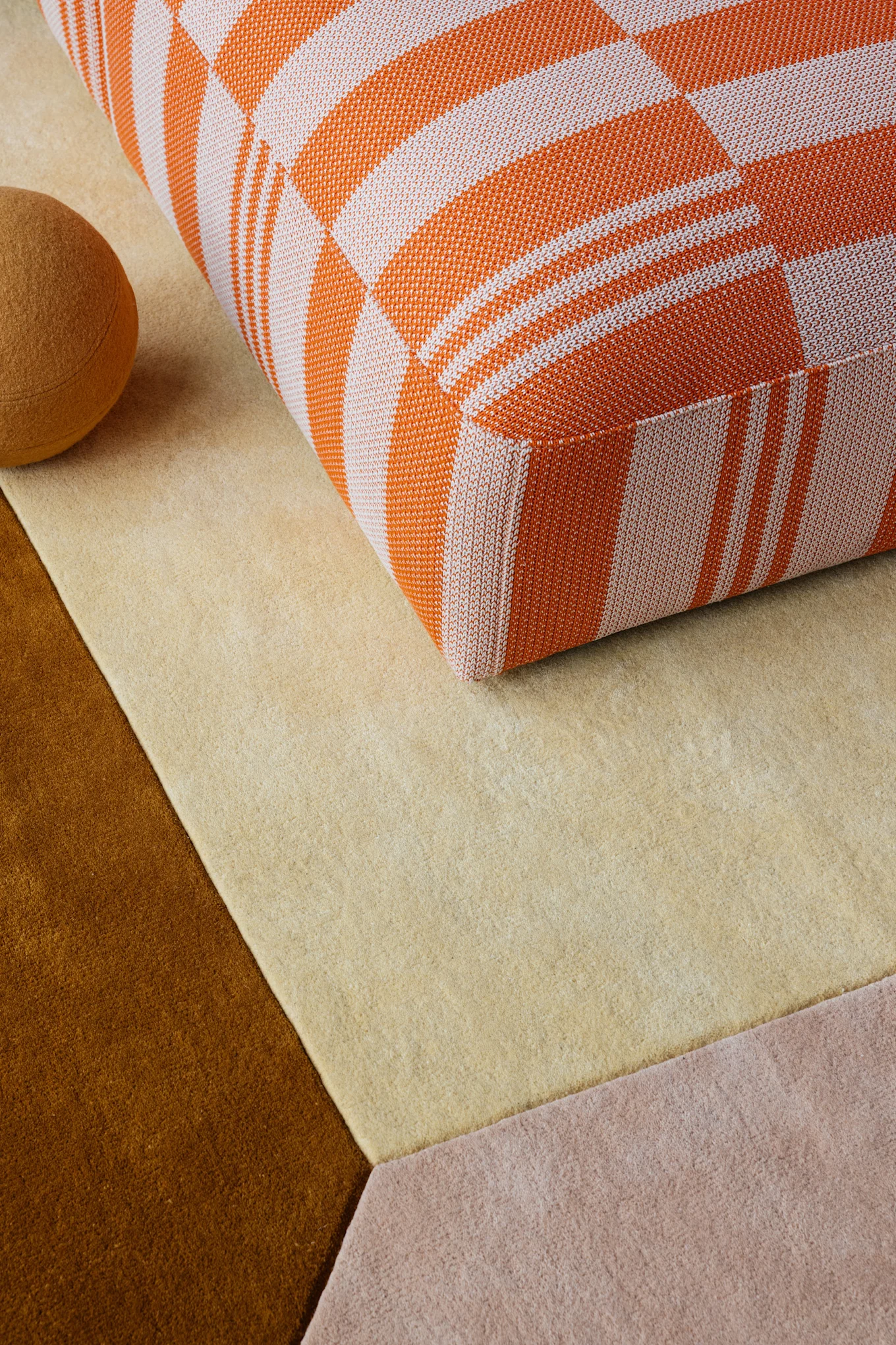 Rutig orange och vit kudde på en geometrisk matta med ljusbeige, bruna och rosa sektioner. En brun sfär finns i närheten.