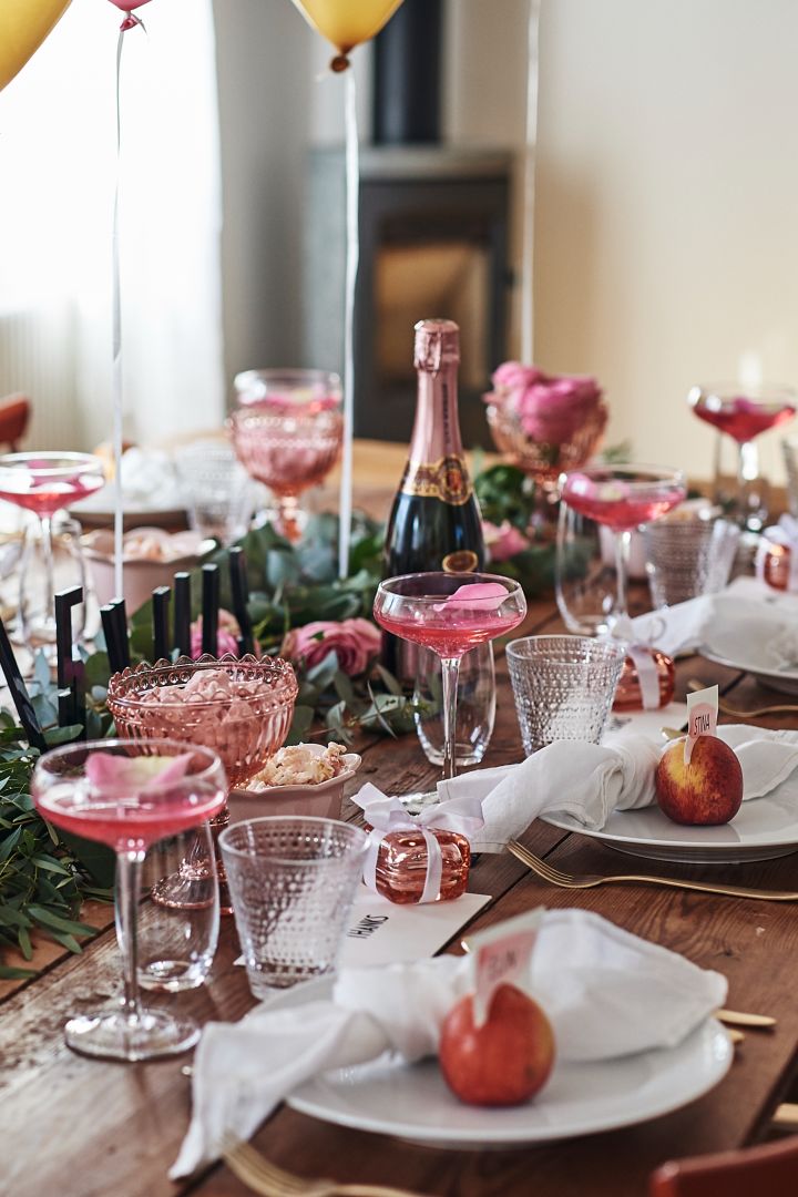 En romantisk och rustik festdukning i rosa toner med Swedish Grace tallrik snö uppdukat på träbordet med cocktailglas och guldbestick. 