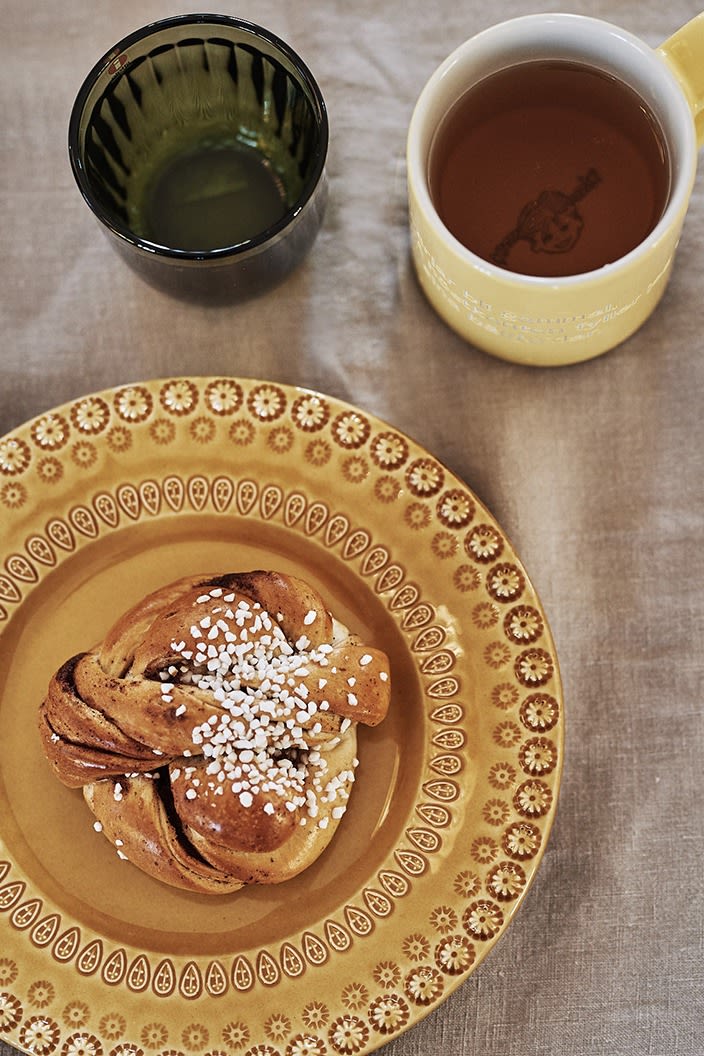 Fikat står framme med kanelbulle på gul assiett från PotteryJo, saft ur Design House Stockholms Astrid Lindgren mugg samt glasen Raami från Iittala.