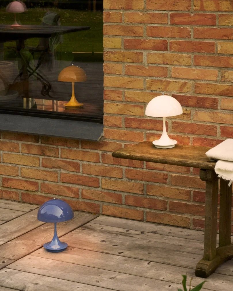 Tre svampformade lampor i vitt, blått och gult på en träterrass vid en tegelvägg och ett fönster.