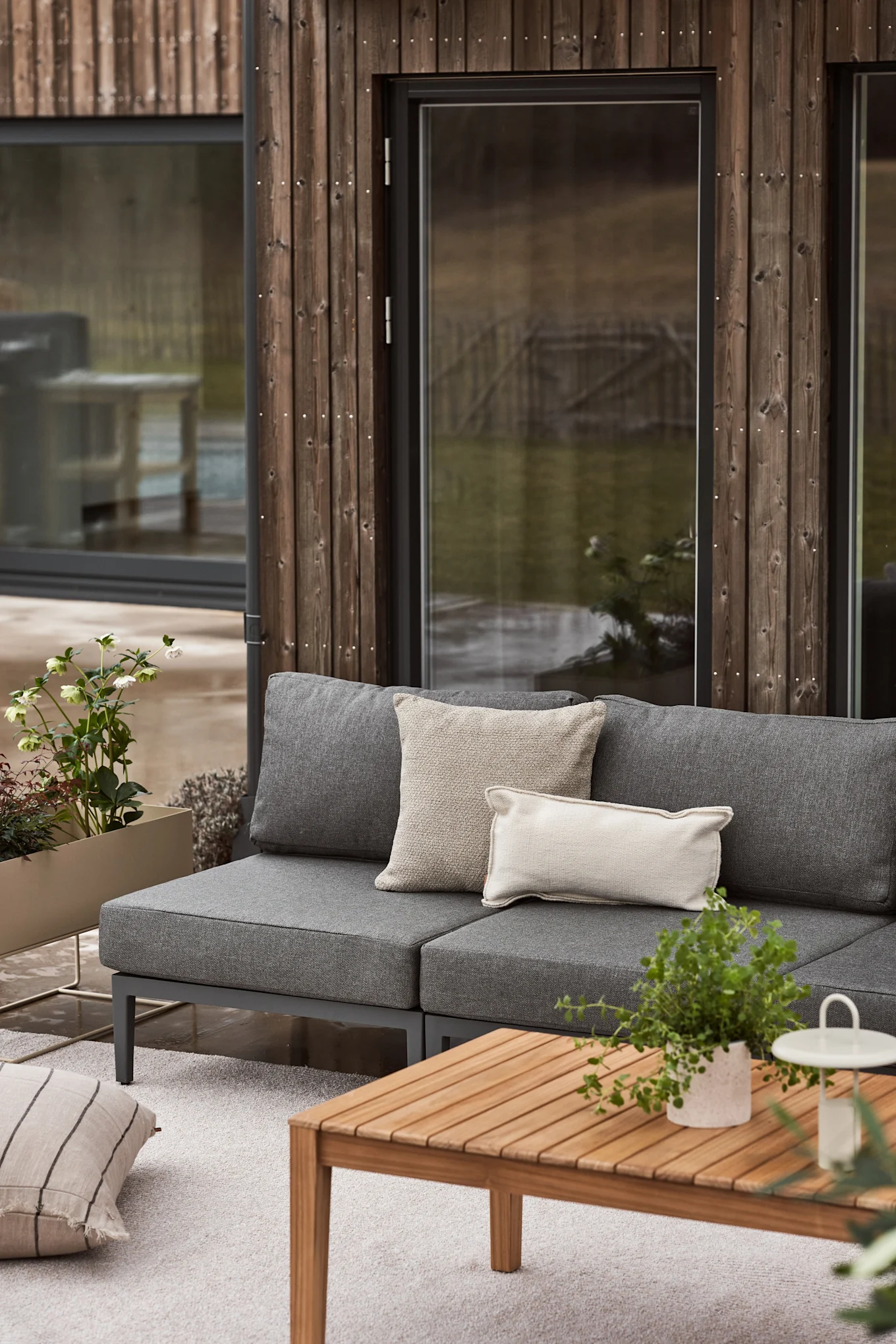 Ett utomhus vardagsrum med en grå modulsoffa, ljusa kuddar, ett soffbord i trä, en ljus matta och planteringskärl med grönska mot en modern träpanelförsedd byggnad. Inspiration till din balkong 2026.