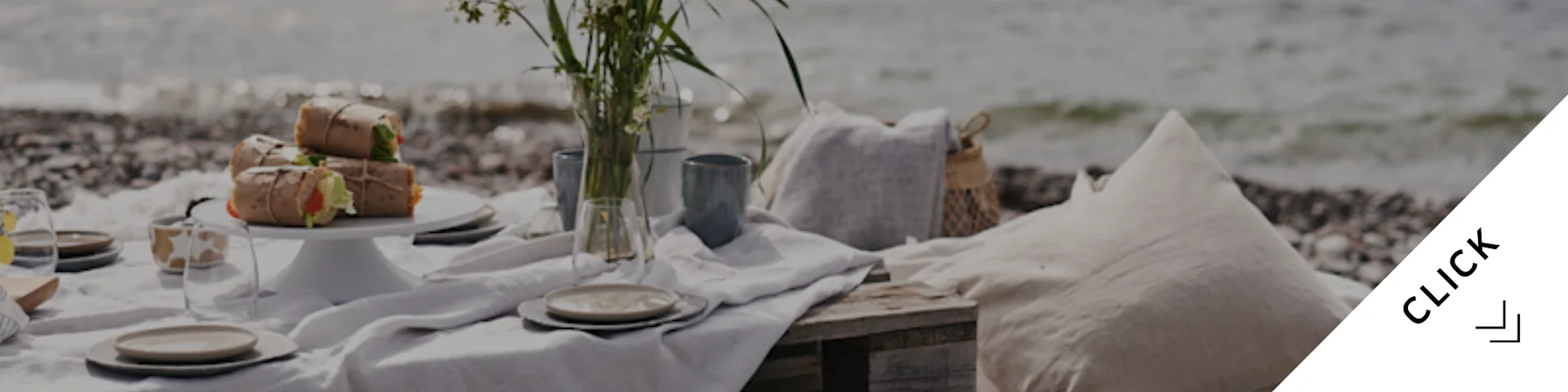 En fridfull strandpicknick med inslagen mat, servis, en vas med blommor och kuddar på en stenstrand vid havet.