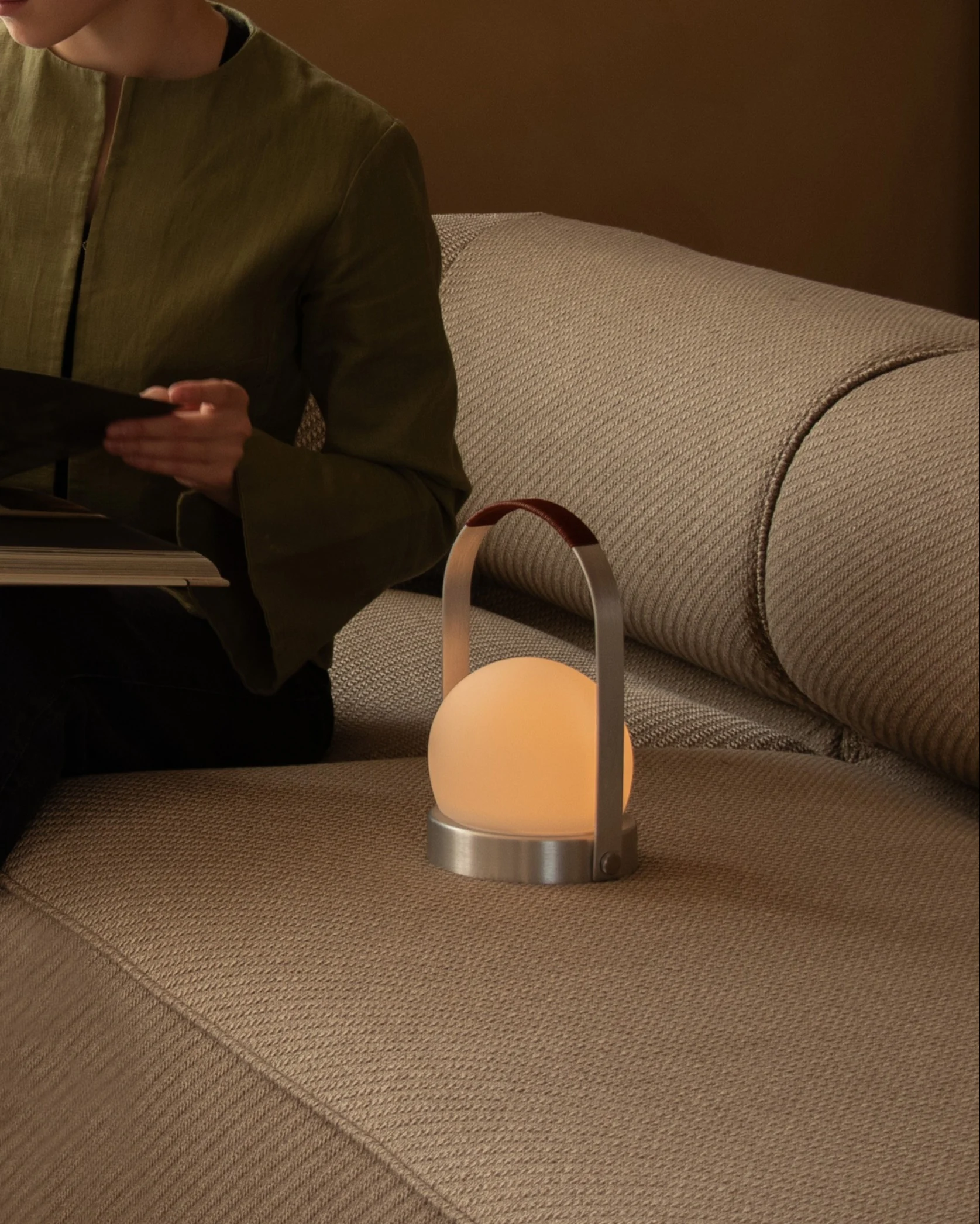 En person läser en bok på en beige soffa bredvid en glödande sfärisk bärbar lampa med läderhandtag.