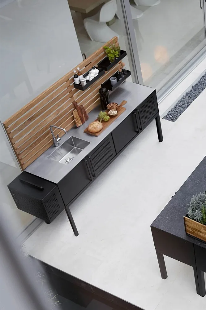 Modernt utekök med diskho i rostfritt stål, svarta skåp, bakpanel i trä och hyllor med kryddor och mat.