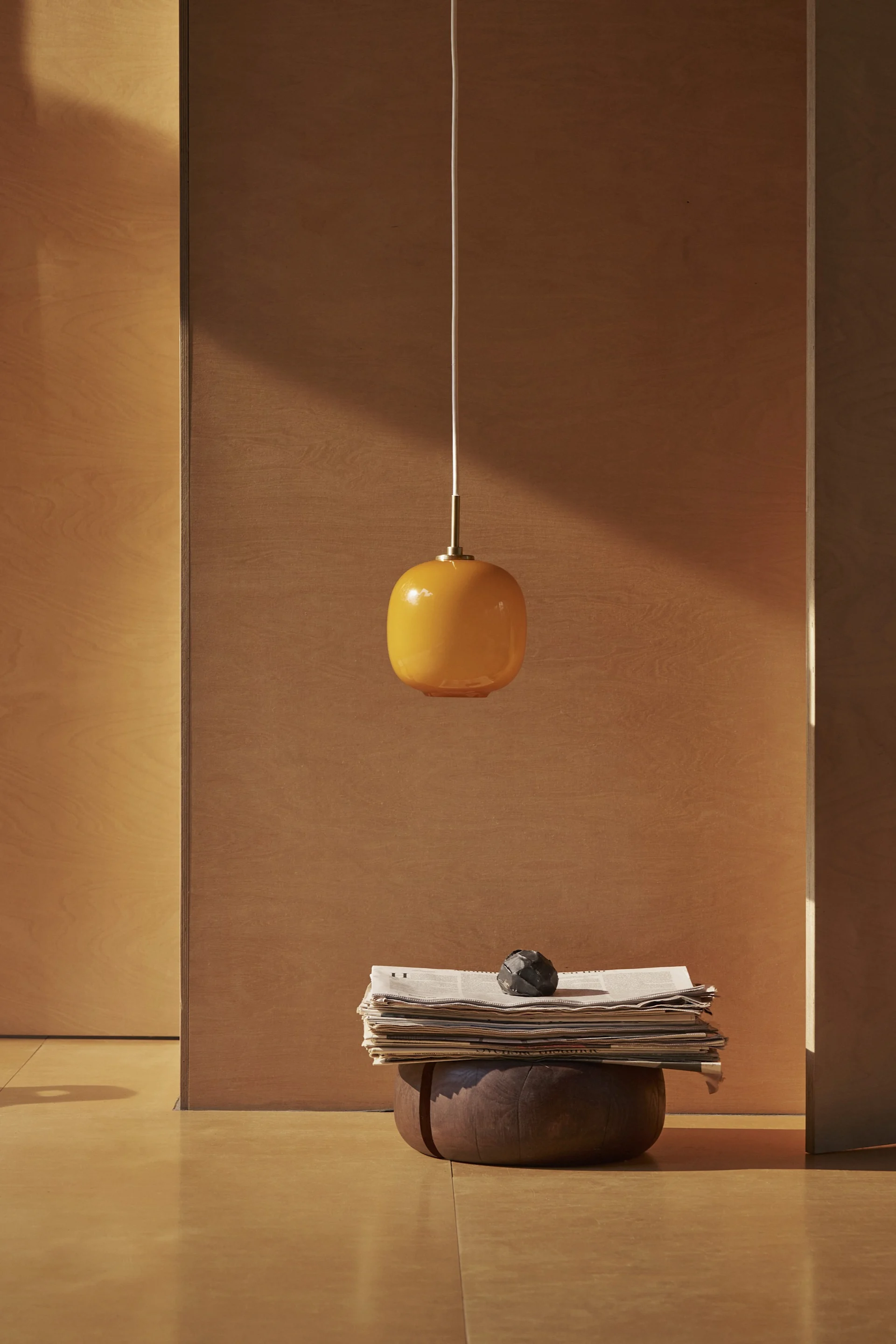 En orange oval pendellampa hänger över en hög med tidningar och ett mörkt dekorativt föremål på en träbas i ett minimalistiskt rum.