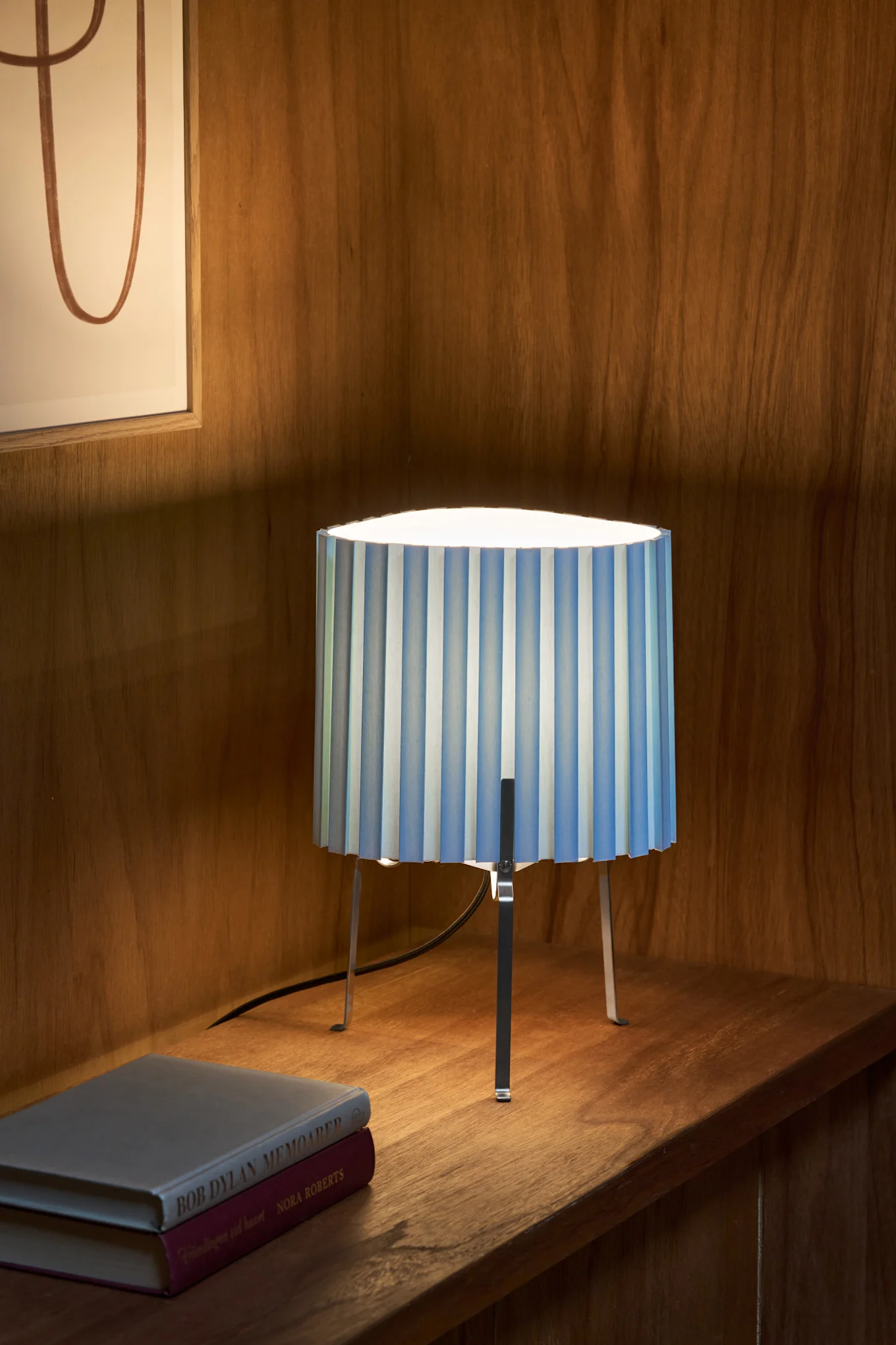 En bordslampa med blå- och vitrandig plisserad skärm och metallben, stående på en trähylla bredvid två böcker.