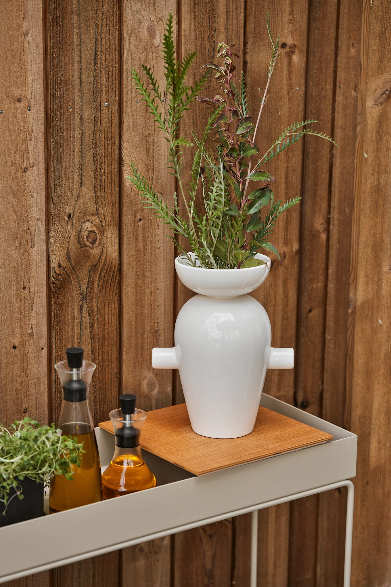 Vit vas med bladverk, två oljeflaskor och en örtkruka på en grå hylla mot ett trästaket. Bli inspirerad att inreda din balkong 2026.