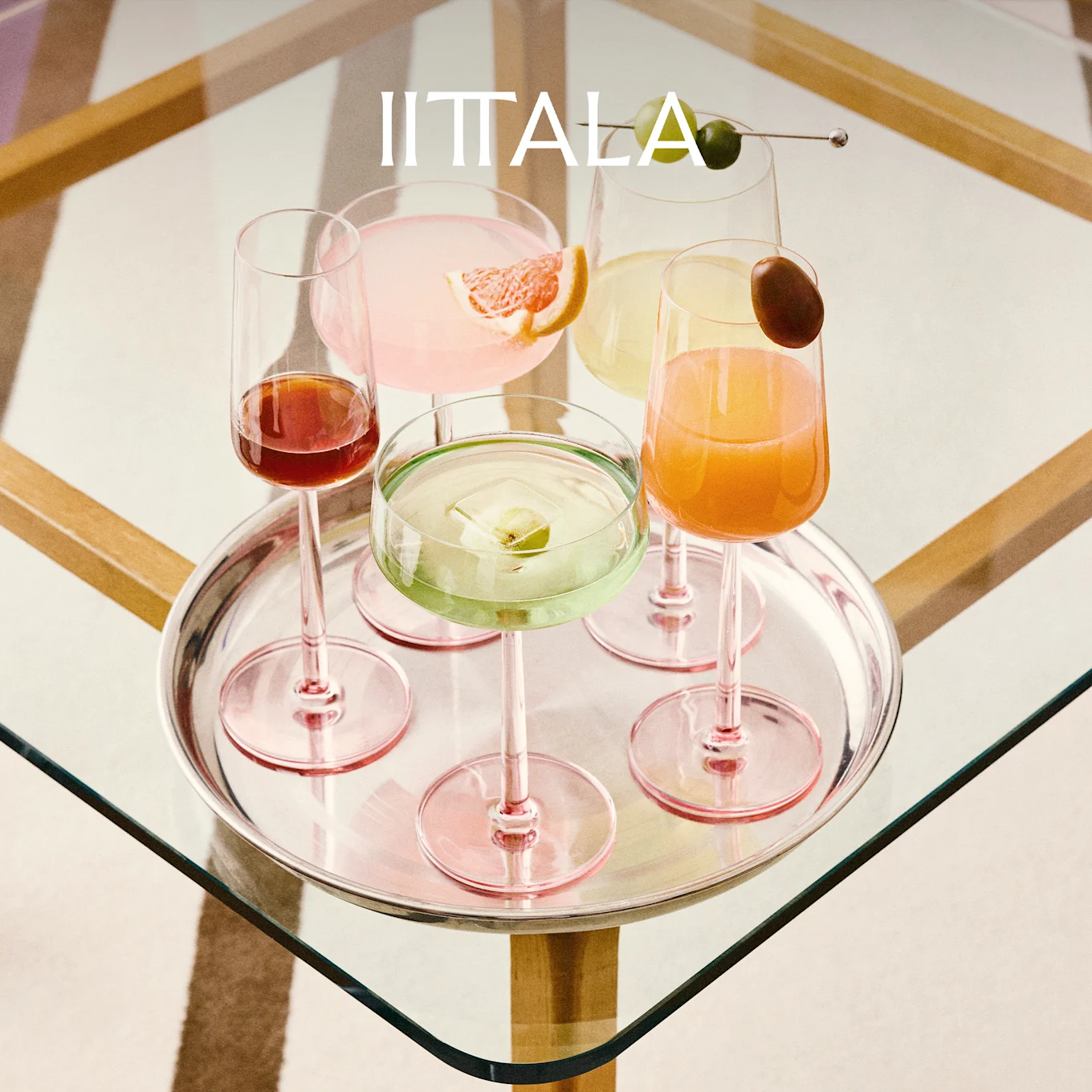 IITTALA-logotyp ovanför en silverbricka med fem färgglada cocktails i rosa-stammade glas på ett glasbord.