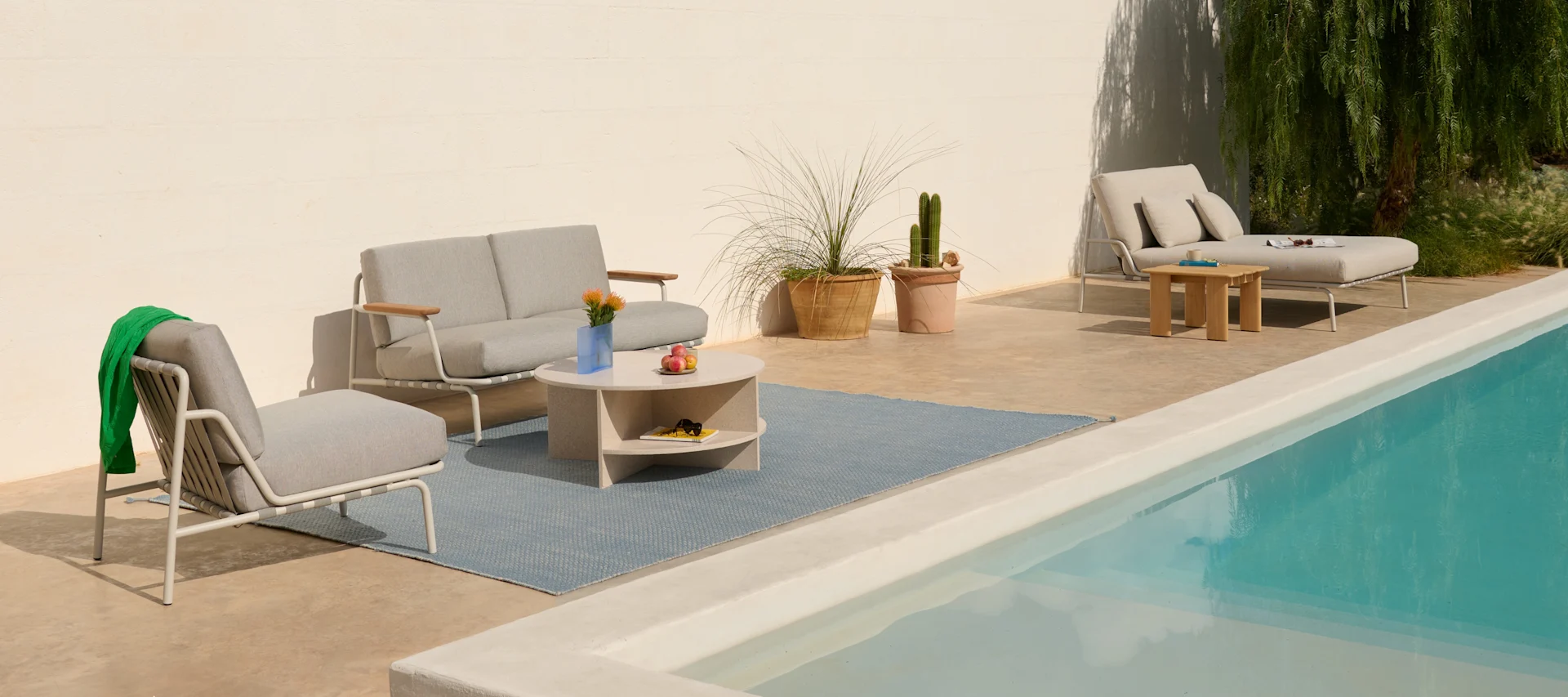 Solig uteplats med ljusgrå utemöbler, blå matta, krukväxter och pool.