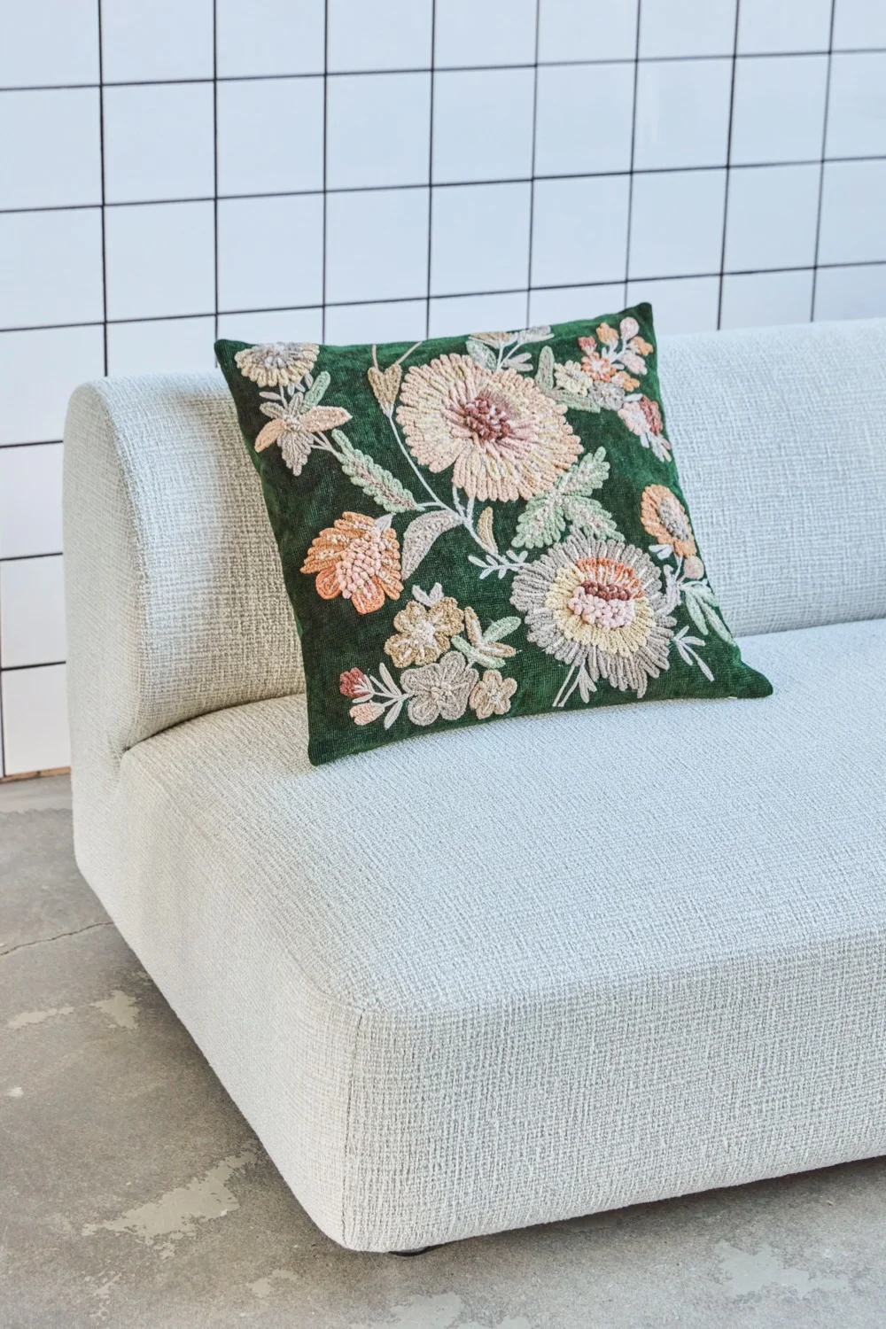 En grön kudde med broderade blommor från House Doctor ligger i en vit soffa med en vit kakelvägg i bakgrunden.