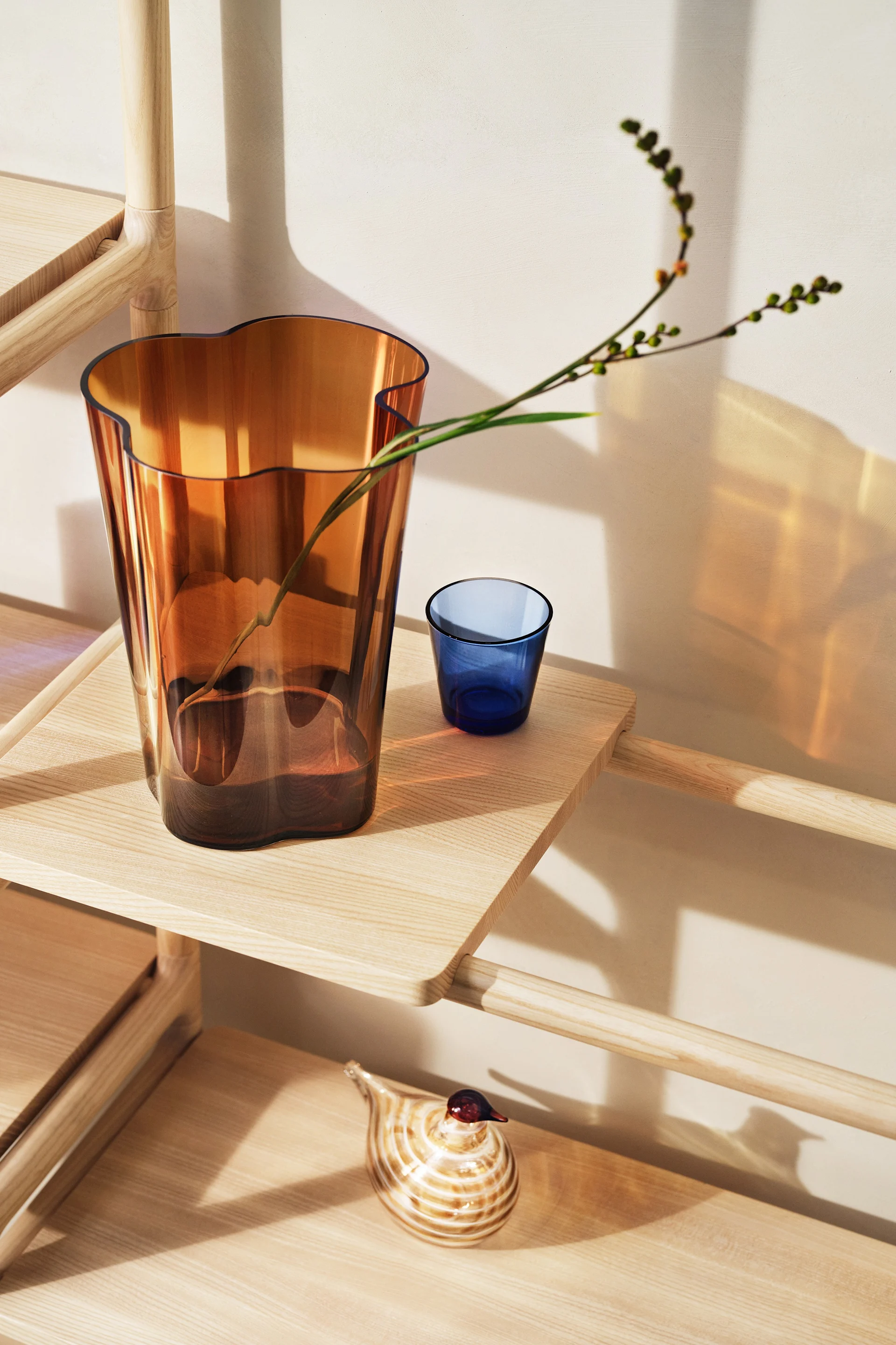 Bärnstensfärgad glasvas med vågig topp och en gren, blått glas och randig glasfågel på en ljus trähylla.
