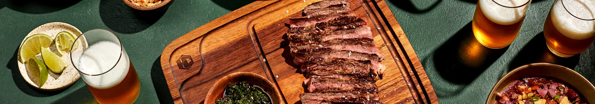 Skivad grillad biff på träbräda med chimichurri, jordnötter, chili och två öl med lime på mörkgrön yta.