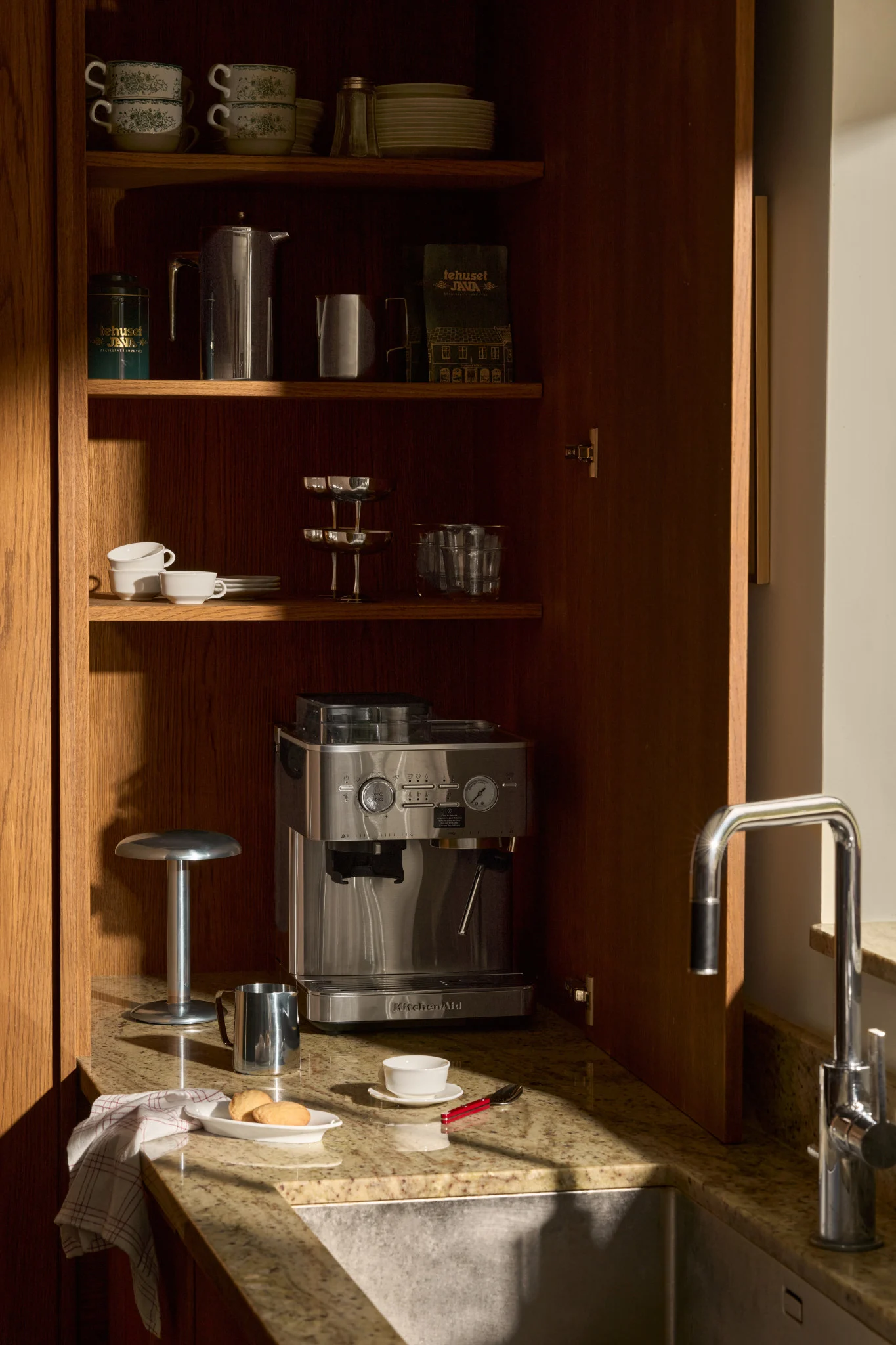 Ett öppet köksskåp i trä med espressomaskin, kaffetillbehör, staplade muggar och glas på hyllor, bredvid en diskho.