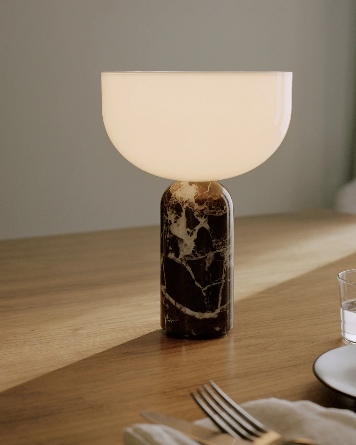 Bordslampa med en cylindrisk mörk marmorbas med vita ådringar och en halvcirkelformad vit glasskärm som lyser, placerad på ett träbord med bestick och ett glas.