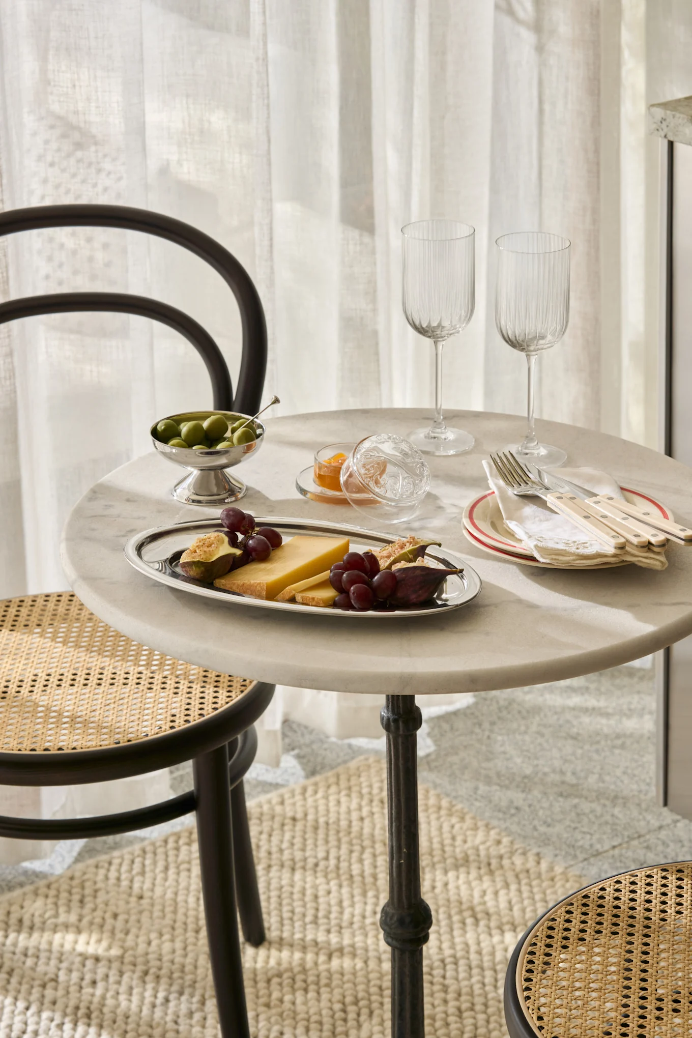 Ett marmorbord dukat med ost, fikon, vindruvor, oliver, sylt, glas och bestick, med rottingstolar.