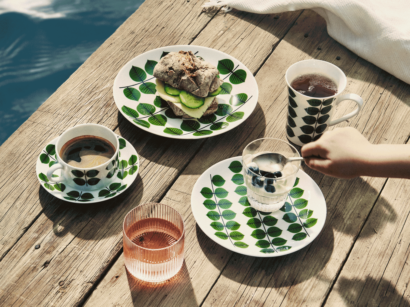Frukost på träbrygga: Bladmönstrade tallrikar, smörgås, kaffe, te, yoghurt med blåbär och rosa dryck.