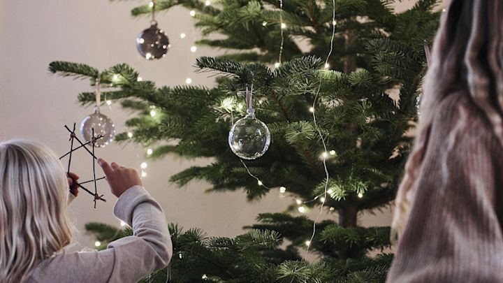 Julgranskulor i glas från Iittala hänger i granen medan barn sätter dit egna julstjärnor av pinnar som de pysslat fram. 