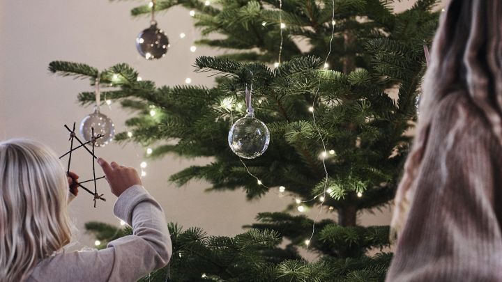 Julgranskulor i glas från Iittala hänger i granen medan barn sätter dit egna julstjärnor av pinnar som de pysslat fram. 