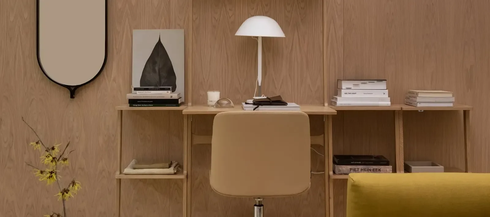 Ett träskrivbord med hyllor, en beige kontorsstol, vit lampa, böcker och en svart spegel mot en träpanelvägg.