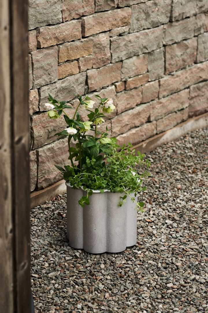 Gråsippa kruka från Byarums Bruk är en riktig designklassiker bland stora krukor för uteplatsen. Krukan är tillverkad i metall och har en vackert veckad design. Här placerad mot stenvägg, planterad med både grönska och blommor.