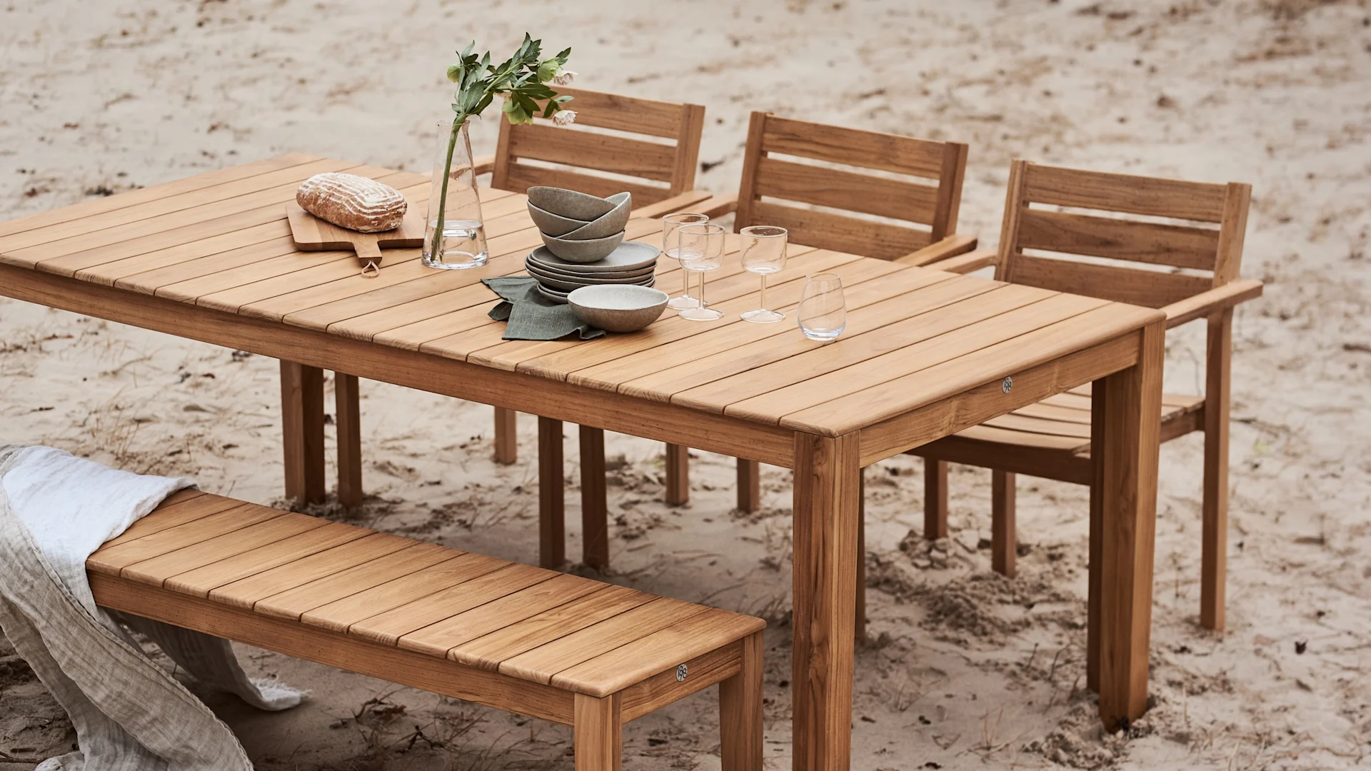 Träutematsalsbord dukat med porslin, glas, en vas med blommor och bröd, omgivet av stolar och en bänk på sand.